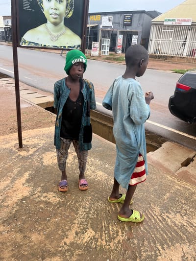 The image shows two young boys standing on a sidewalk in Sabon Gida, Nigeria. The boy on the left is wearing a light blue, long-sleeved garment and red and white striped shorts. He is looking upwards and to the left, with his right hand partially raised. He is wearing bright green slip-on shoes.

The boy on the right is wearing a green beanie, a dark vest with visible holes, and patterned pants. He is also wearing colorful slip-on sandals. He is looking downwards and slightly to the right.

Behind the boys, there is a paved road and buildings. One building has signs for "NKMAKEL PHARMACY & STORE" and "CLASSES". Another building has signs for "PRINTING", "A3, A4 PHOTOCOPY", and "CODING CLASSES". A large billboard featuring a woman's face and text describing services like "Frames", "Photobooks", "Branding", "Trainnings", "Coverages", and "Make up and more" is also visible on the left side of the frame. A dark car is parked near the sidewalk. The ground is a rough, textured concrete. The lighting suggests it is daytime. The overall scene appears to be a street in a commercial area.