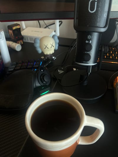 This image captures a close-up, top-down view of a personal desk setup, likely a gaming or content creation station. The scene is indoors, featuring various electronic devices and personal items arranged on a dark, possibly textured or carbon-fiber patterned surface.

The most prominent subjects include a black microphone with a pop filter, standing on a round base. The microphone features control knobs and buttons, with visible text indicating "ECHO" and "VOLUME," along with icons for light settings and microphone gain/mute. A partial "O" suggests the brand HyperX. To its right, a white and brown (or copper-toned) ceramic mug, filled with a dark liquid resembling coffee, sits on the desk.

To the left of the microphone, partially obscured, is a white electronic device, possibly a Wi-Fi router, with visible vents. Next to it stands a light-colored KAWS Companion figurine, characterized by its stylized head and "X"s for eyes, with grey puffy elements. Below these items, a miniature arcade cabinet is visible.

In the foreground, a gaming keypad with an integrated wrist rest is positioned. This keypad features multiple keys with colorful LED backlighting and a joystick/D-pad control. Visible text on the keys includes numbers like "20." Cables connect the microphone and other devices. A monitor or screen is partially visible on the far left edge of the frame.

The lighting suggests an indoor environment, possibly ambient room light or artificial light, creating soft shadows. There are no direct visual cues in the image that specifically indicate the location is Mérida, Mexico; the setup appears generic for a modern desk. No direct human activity is depicted, but the arrangement of items implies a user engaged in tasks like gaming, audio recording, or general computer use.