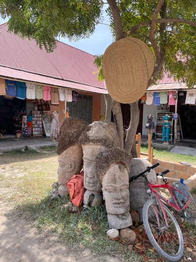A red bicycle rests atop a collection of weathered, carved wooden heads. Next to them, a shop displays an array of bags and souvenirs under a corrugated red roof. A woven, oval-shaped mat hangs from a tree branch near the shop. Vegetation surrounds the scene, which is situated alongside a sandy path.