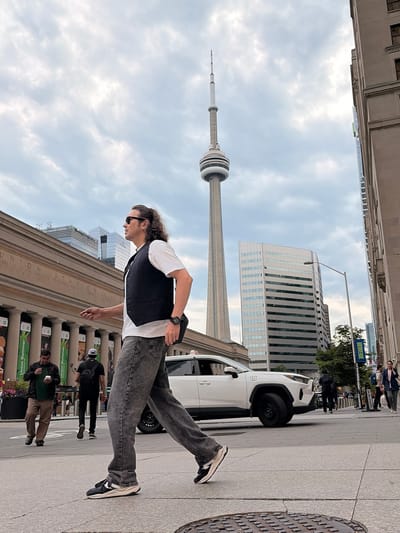 The image captures a street scene in Toronto, Canada, with the CN Tower prominently visible in the background. In the foreground, a man is walking towards the right of the frame, wearing a white t-shirt, a dark vest, and dark, baggy jeans. He has dark hair pulled back and is wearing sunglasses and a watch. A white SUV, possibly a taxi or ride-share vehicle, is parked behind him, partially obscuring his lower body. To the left of the main subject, the facade of Union Station is visible, with several people walking on the sidewalk in front of it. One man in a green shirt and brown pants is looking at his phone, and another person in a black t-shirt and backpack is walking away from the camera. The environment appears to be a busy urban area with tall buildings and a cloudy sky.