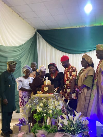 A group of people are gathered around a table. On the table, there are flowers and gifts. The people are dressed in traditional clothing. The environment appears to be a venue decorated with green and white fabrics. The location is Vom, Nigeria.