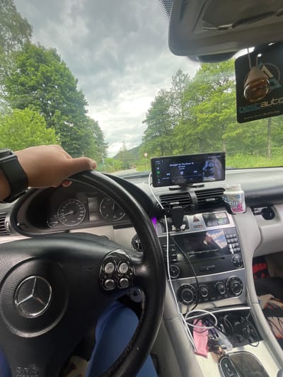 The image captures the interior of a Mercedes-Benz vehicle, viewed from the driver's perspective. A left hand, adorned with a black watch, grips the leather-wrapped steering wheel, which features the iconic Mercedes-Benz logo. The dashboard displays a digital speedometer and other instruments. Mounted on the center console is an aftermarket touchscreen infotainment system, currently playing music by 50 Cent. Next to it, a can of "Orbit White" gum and a car air freshener are visible. The car is driving on a road surrounded by lush green trees, with a cloudy sky overhead, suggesting a rural or mountainous setting. The location is identified as Elhovets, Bulgaria.