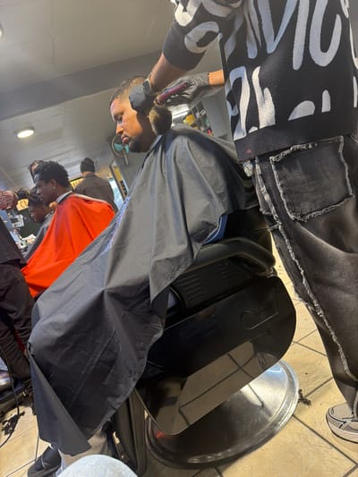 Inside a Blouberg, South Africa barbershop, a barber with a black glove is cutting the hair of a client, who is draped in a black barber cape. Another patron with an orange barber cape awaits their turn. The scene is set on tile flooring. A silver barber tray is visible. The barber is wearing a black and white patterned sweater and distressed black jeans.