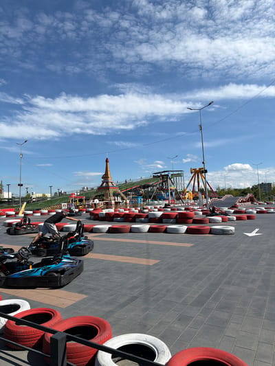 This is an eye-level, outdoor, full shot of a go-kart track. A man in a grey t-shirt and shorts is pushing a go-kart. Several other go-karts are parked or in use on the track. The track is bordered by stacks of red and white tires. In the background, there is an amusement park with a Ferris wheel, a roller coaster, and a replica of the Eiffel Tower. The sky is blue with scattered white clouds. The location is the go-kart track in Tohatin, Moldova.