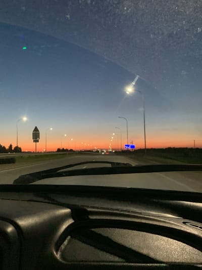 The image shows a view from inside a car, looking out through the windshield. It is dusk, with a gradient of colors in the sky ranging from deep blue at the top to orange and pink near the horizon. A highway stretches into the distance, lined with streetlights. On the left side of the road, there are trees and a road sign. On the right side, more streetlights and trees are visible. The dashboard of the car is in the foreground, partially obscuring the view. There are no people visible in the image, and no specific actions or interactions are taking place. The environment appears to be a suburban or rural highway setting. The image was taken in Melekes, Russia, likely at sunset or sunrise due to the sky colors.