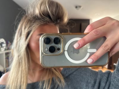 A woman's face is partially obscured by her iPhone, held up in a selfie. The phone has a clear case with gold trim and a white MagSafe ring. Her hair is blonde, styled in a messy updo with loose strands framing her face. She wears a gray, off-the-shoulder top. A bookshelf, decorated with colorful items, is visible in the blurred background. A smoke detector is on the ceiling.