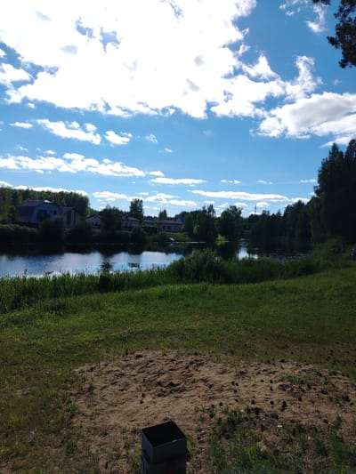 The image depicts a tranquil waterscape with a residential area in the background. A calm body of water, reflecting the bright blue sky dotted with fluffy white clouds, occupies the mid-ground. The water is bordered by lush green grass and reeds on the foreground bank. On the opposite bank, a cluster of houses with various rooflines and architectural styles are nestled amongst dense green trees. The houses appear to be part of a rural or suburban settlement. The foreground consists of a patch of sandy ground with scattered pine cones and some green grass. A dark metal object, possibly a grill or trash receptacle, is visible in the lower right corner of the sandy area. The overall impression is one of a peaceful summer day in a natural setting with residential development. The location is confirmed to be Stroitel', Russia.