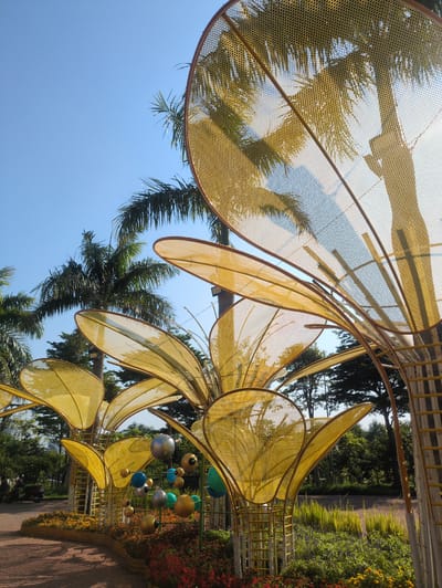 The image presents a tilted view of a public art installation in Tuy Hòa, Vietnam. The installation features large, stylized yellow floral shapes constructed from a mesh material, resembling petals. The structure is partially framed against a clear blue sky and palm trees. To the right, a walkway is lined with vegetation, including flowering shrubs and a variety of plants. Spherical ornaments in colors of blue, green, silver, and gold are suspended from the structure. The scene suggests a sunny day.