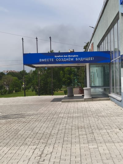 The image shows the entrance to the Komsomolsk-on-Amur Regional Youth Center. A large blue banner stretches across the entrance, with Russian text that translates to "Together we are creating the future!". The banner is supported by two dark poles with decorative finials. The building itself is light blue with large glass windows. In front of the building, a paved walkway made of light gray bricks leads to the entrance. Two cylindrical planters, also light gray, are placed on either side of the walkway. Beyond the entrance, a park-like area with green grass and trees is visible. The sky is overcast with some clouds. The scene is brightly lit by natural daylight.