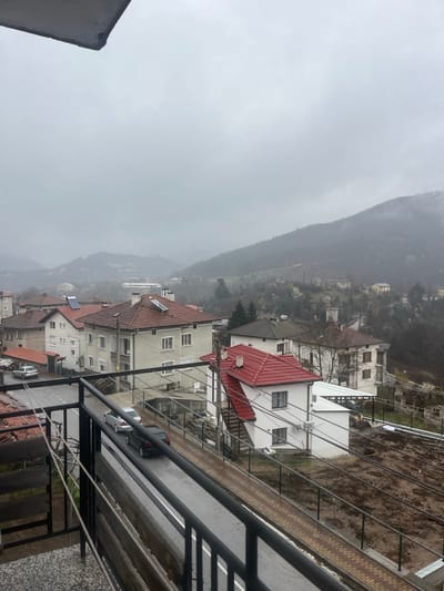 This image depicts a view from a high vantage point, likely a balcony, overlooking a residential area in what appears to be Srednogortsi, Bulgaria. The sky is overcast and foggy obscuring the mountain range in the background. The houses are built close together, some with red-tiled roofs, others with white walls and red roofs, creating a layered effect down the slope. A street runs parallel to the balcony, with at least two cars visible: one dark-colored, and the other silver-colored parked closely together. The balcony features metal railings, and wires run overhead, possibly for electricity or other utilities. Overall, the scene conveys a sense of density and subdued weather conditions.