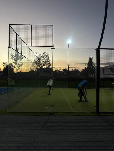 Two padel players are playing on a court at sunset. One player in a blue shirt is in mid-swing, while another in a white jacket stands ready. The court is enclosed by glass walls and wire mesh fencing, illuminated by an overhead light. Trees are visible in the background, silhouetted against the colorful sky. A paved walkway runs along one side of the court.