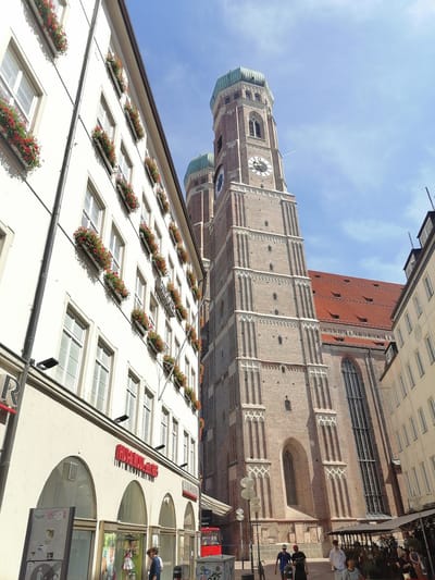 This low-angle shot captures a sunlit street scene in Munich, Germany, dominated by the impressive architecture of a Gothic cathedral. The cathedral's tall brick tower with a green dome and a clock face rises into a clear blue sky with wisps of clouds. Adjacent to the cathedral, a white building features rows of windows adorned with flower boxes overflowing with red blooms. The building also displays red signage with text that includes "HOHENZOLLERN" and appears to be a shop or restaurant.
Below the white building, the street level reveals storefronts with large windows displaying mannequins. A red public bus is visible in the distance. Several people are walking on the sidewalk, some in casual attire. The sunlight casts distinct shadows, indicating it is likely daytime, possibly late morning or early afternoon. The atmosphere appears to be calm and pleasant, with the bright sky and flourishing flowers suggesting good weather. The scene conveys a typical urban environment with historical architecture and daily life unfolding.
