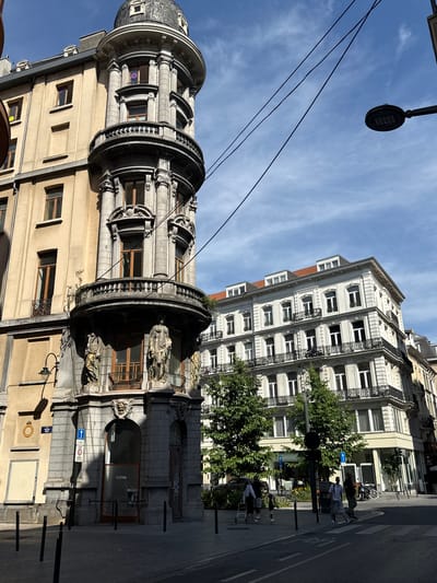 This is a vertical, eye-level shot of a street in Brussels, Belgium, on a bright, sunny day with some wispy clouds in the sky. The image is dominated by two ornate buildings. The foreground building on the left is characterized by a curved, domed turret with intricate stonework and decorative balconies. The building to its right is a taller, more rectangular structure with multiple floors, each featuring rows of windows with decorative wrought-iron balconies.
Several people are visible on the sidewalk and crossing the street, suggesting a normal daytime scene. Some are walking, while others appear to be standing and conversing. One person is near a bicycle. The scene is urban, with streetlights and power lines also visible. The sunlight creates distinct shadows, indicating it is likely mid-morning or early afternoon.
Notable details include the architectural styles of the buildings, which suggest an older European city. There are also signs visible, one of which reads "TATTOO" on a shop window. Another sign appears to be a traffic sign with a blue arrow and a circle with a number 30 inside, likely indicating a speed limit. The overall mood is lively and ordinary for a city street.