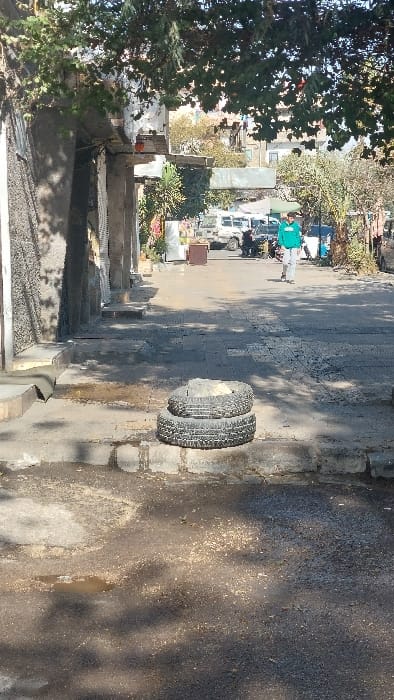 The image depicts a daytime street scene within Al-Qaboun Municipality, Syria. In the immediate foreground, a dark asphalt surface with scattered light debris and a small puddle transitions to a raised, light-colored concrete curb. Two heavily worn vehicle tires are stacked on the curb-side edge of the adjacent paved pedestrian area, with light-colored material visible on the upper tire. Shadows from overhead foliage are cast across the foreground ground and paved areas.

To the left, a row of weathered buildings with light-colored facades and visible steps leading to entrances lines the pedestrian path. Overhanging tree branches with green leaves partially obscure the upper left section.

The midground features a paved street or alley extending into the distance. A lone male individual is walking away from the viewer on the right side of this street, wearing a light teal or green hooded sweatshirt and light-colored trousers. Further down the street, multiple parked vehicles are visible, including at least two white SUVs or jeeps. Green foliage from trees and bushes flanks the right side of the street and appears throughout the background.

The background consists of additional buildings and dense greenery under a bright, clear sky. The overall scene is illuminated by strong sunlight, producing distinct shadows.