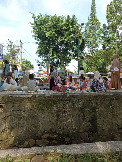 This outdoor image depicts a lively community gathering or market event in Mataram, Indonesia, bathed in bright daylight under a clear, blue sky. The foreground is dominated by a rough-textured concrete and stone wall, on which numerous individuals are casually seated.
The primary subjects are several women, mostly wearing hijabs, and young children. Many are seated along the wall, with children appearing to be engaged in an activity or eating snacks from small plates in front of them. One woman in a grey hijab and black top is seated prominently on the left, while another in a floral top is on the right. A young boy in a grey shirt is seated centrally on the wall, facing away from the camera. Other adults are seen standing or sitting further back, interacting around several white tables that hold items like bottled drinks, suggesting market stalls or activity stations.
In the mid-ground and background, colorful market umbrellas (red, blue, yellow) are visible, providing shade over other tables. The scene is lush with natural greenery, featuring dense trees with bright green foliage, and a distinctive tall, slender tree on the right. A decorative street light pole stands centrally in the mid-ground. On the far left, utility poles and power lines are visible, along with a partial white banner displaying the text "AN".
The weather is clear and sunny, indicative of morning or early afternoon. The overall mood appears relaxed and social, characteristic of an open-air community event or informal market.