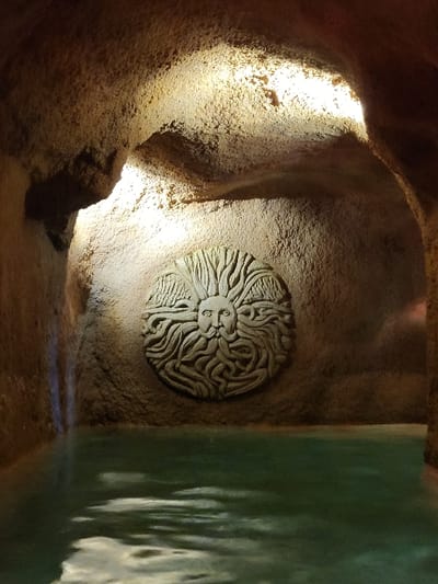 A textured, circular sun sculpture adorns a wall within a cave-like setting. Light illuminates the wall around the sculpture from the left side. To the right, a glass structure allows light to shine through and creates a blurry, greenish image. The cave walls and floor are a mix of sandy and brown hues. The scene appears to be indoors.