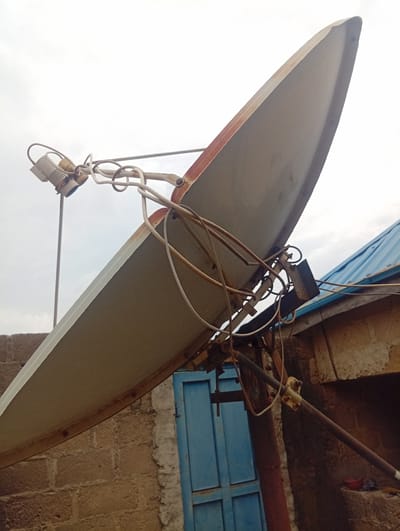 A satellite dish, weathered with rust along its rim, is angled upwards against a bright, overcast sky. The dish is mounted on a metal pole with visible wires and hardware connecting to a receiver unit at the focal point. A second, smaller dish or antenna is attached to the main mount.
Behind the satellite dish, a building constructed of rough, tan-colored bricks is partially visible. A section of a blue metal door or gate is also in the frame, to the left of the building. To the right, a section of a blue corrugated metal roof juts out from the building. The overall scene suggests an outdoor setting, likely residential, given the building materials and the presence of a satellite dish for television reception. The cloudy sky indicates it could be daytime, but the diffused light suggests either early morning, late afternoon, or a generally overcast day. There is no visible text in the image. The location context provided places this scene in Gwada, Nigeria.