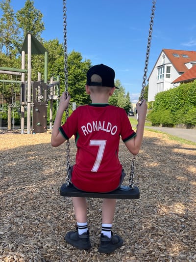 A young boy, viewed from behind, sits on a swing in a playground. He is wearing a red soccer jersey with the name "RONALDO" and the number "7" printed on the back in white. He also wears a black baseball cap, grey shorts, black sneakers with white striped socks. The playground has a wood chip ground cover. In the background, a wooden climbing structure and trees are visible. To the right, a white house with a red-tiled roof and green bushes are present. The scene is set on a clear, sunny day. The location is Sachsenheim, Germany.