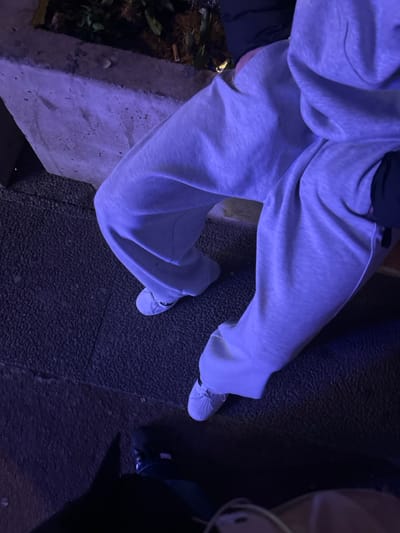 A downward shot depicts a person wearing light gray sweatpants and white sneakers. The clothing appears baggy. They stand on a dark, textured surface. To the left is a concrete structure with greenery and possible cigarette butts. In the right foreground are partial views of other people, including one with dark sneakers. The image is dominated by shades of blue.