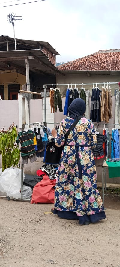 A woman wearing a floral dress and a hijab stands in front of a clothing rack at a market in Kecamatan Kutawaringin, Indonesia. The rack is filled with assorted children's clothes, hanging on metal hangers.  A green plastic table sits to the right of the rack.  There are various objects surrounding the table and clothing rack, including red, black, and grey bags. Green beans hang from a metal pole in the background. There is a building in the background, with a brown tile roof. The pavement is dusty, with a dirt-like surface.  The woman is looking down at the clothing on the rack.