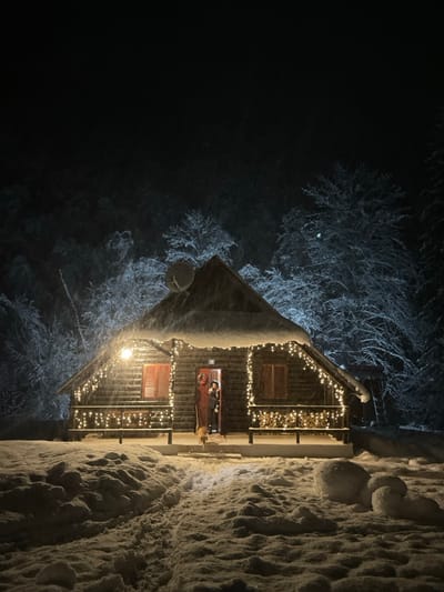 The image depicts a charming wooden cabin or lodge, festively adorned with warm string lights, set against a dark, snowy night in Lugovi, Montenegro. The original image is rotated 90 degrees clockwise; when viewed correctly, the cabin stands upright amidst a winter wonderland.
The cabin, constructed from dark timber, is blanketed in thick snow that covers its roof, balcony railings, and the surrounding ground. Numerous small, glowing yellow lights are strung along the cabin's exterior, illuminating the porch area and creating a cozy, inviting atmosphere. Two windows with closed wooden shutters are visible on the facade.
On the illuminated porch, a person is seen standing near an open doorway, wearing a dark coat and hat, suggesting cold weather. Their face is indistinct due to the distance and lighting. Adjacent to the person, a small, light-colored dog is also visible. A red wreath, likely festive, hangs near the entrance. Stacked logs of wood are piled on the porch, and a satellite dish is mounted on the cabin's left side.
The scene is dominated by a heavy snowfall, with snow-laden trees appearing ghostly white in the dark background. The ground is covered in deep, textured snow. The overall impression is one of a tranquil, cold winter evening, possibly during the holiday season, with the warm lights of the cabin providing a beacon of comfort. No visible text is present in the image.