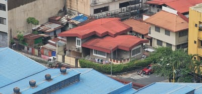 The image depicts a dense urban landscape in Makati, Philippines, characterized by a mix of residential structures and commercial buildings. In the foreground, a portion of a large blue-roofed industrial or commercial building is visible, featuring several industrial ventilation units.

Behind this, a cluster of older, somewhat dilapidated houses with corrugated metal roofs, some rusted, are tightly packed. A few small trees are interspersed among these structures. Adjacent to these, a more substantial concrete house with a red tiled roof stands out. This house has a balcony, windows with blinds, and an air conditioning unit. Laundry is hung out to dry on a fence in front of it.

Further back and to the right, a cream-colored building with multiple windows and a reddish-brown roof is situated. Next to it, a tall yellow building with visible windows and air conditioning units suggests a commercial or office space.

A red car and a motorcycle are visible on the street, crossing a pedestrian lane. The street is paved and has visible markings. Lush green vegetation, including trees and bushes, surrounds some of the properties, creating a contrast with the man-made structures. The overall impression is a layered urban environment with varying degrees of development and upkeep.