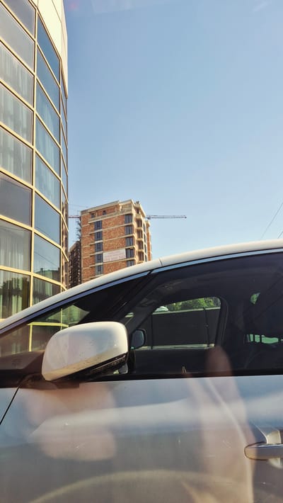This is a photograph taken from inside a vehicle in Ialoveni, Moldova. The image captures a construction site with a building under construction, identifiable by its brick facade and scaffolding. A banner on the building reads "Vanzare Apartamente 060 800 500". A crane is visible extending horizontally in front of the building. To the left of the construction site is a modern building with a glass facade. The foreground of the image shows the interior of the vehicle, including the side mirror and a portion of the car's door and window. The sky is clear and blue, suggesting a sunny day. There are no people visible in the frame.