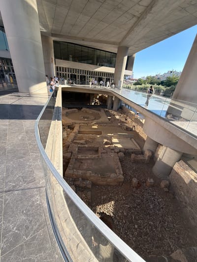 The image depicts an outdoor scene within a modern architectural structure that offers a view of an archaeological excavation site. Several people are visible, some standing and observing the ruins, while others appear to be walking along a glass-enclosed walkway. The walkway has a metal railing, reflecting the surrounding environment.
The archaeological site features ancient stone structures, including walls, foundations, and what appears to be a circular foundation or well. The ground within the excavation is covered with loose stones and dirt.
The setting suggests a museum or historical site with a contemporary design, possibly an extension or viewing platform built over or adjacent to the ruins. The bright sunlight and clear blue sky indicate a daytime scene, likely in pleasant weather. The architecture includes large concrete elements and glass facades.
The people's attire suggests casual wear, suitable for a warm day. Their posture and orientation suggest they are engaged in observing and exploring the site.
The location is identified as Athens, Greece, which aligns with the presence of ancient ruins and the architectural style. The contrast between the modern building and the ancient remnants is a prominent feature. There is no visible text in the image.