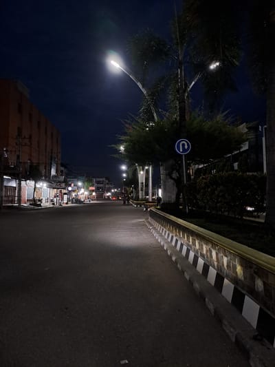 The image depicts a street scene at night in the city of Bireuen, Indonesia. The street is empty and illuminated by streetlights. On the left side of the street, there is a building with several windows and shops on the ground floor. Some of these shops have signs, including one that says "ASC" and another for "Oppo". Further down the street, more buildings and streetlights are visible. On the right side of the street, there is a decorative wall with a pattern of black and white squares, bordering a patch of green vegetation and trees. A traffic sign indicating a U-turn is visible on a pole near the trees. The sky is dark, suggesting it is late at night. There are no people actively engaged in any interaction in the foreground, though a few figures can be seen in the distance. The overall atmosphere is quiet and still.