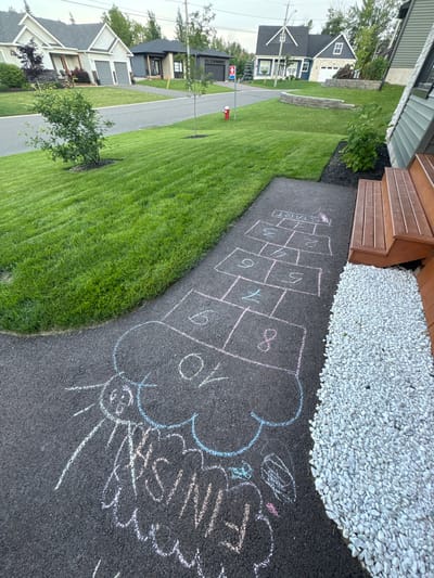 In Dieppe, Canada, on a paved walkway adjacent to a well-maintained green lawn, a hopscotch grid has been drawn with chalk. The grid is marked with pink chalk, numbering squares 1 through 8, with a "START" designation at the top. Below the hopscotch grid, additional chalk drawings are visible, including a cloud shape and what appears to be the word "FINISH" written in cursive. The surrounding environment features residential houses, trees, and a wooden deck with stairs.