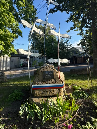 In the city of Kotelniki, Russia, on a sunny day, a monument is prominently displayed. The monument features three white metal sculptures of cranes in flight, mounted on slender poles. One crane is positioned in the foreground, two are in the mid-ground. The base of the monument is a large, rough-hewn stone rock with inscriptions. The top inscription on the rock reads "РУСЬ" (Rus'). Below that, in quotes, is "КРАЕУГОЛЬНЫЙ КАМЕНЬ" (Cornerstone) followed by the date "24.02.2022 г." (24.02.2022). Further down, the word "БЕЛАРУСЬ" (Belarus) is carved. The monument is adorned with a tricolor ribbon in red, white, and blue, positioned horizontally across the rock below the inscription "БЕЛАРУСЬ". At the very bottom, "НОВОРОССИЯ" (Novorossiya) is inscribed on the rock.
The monument is surrounded by a small garden bed with green plants and some pink flowers. To the left of the monument, a bus stop structure with a bench is visible, where two individuals are seated. A grey corrugated metal fence is behind the bus stop. In the background, trees and residential buildings can be seen under a clear blue sky with a few wispy clouds. The sun casts shadows, indicating it is likely late afternoon. The overall scene depicts a roadside memorial with symbolic elements related to national identity and historical events.