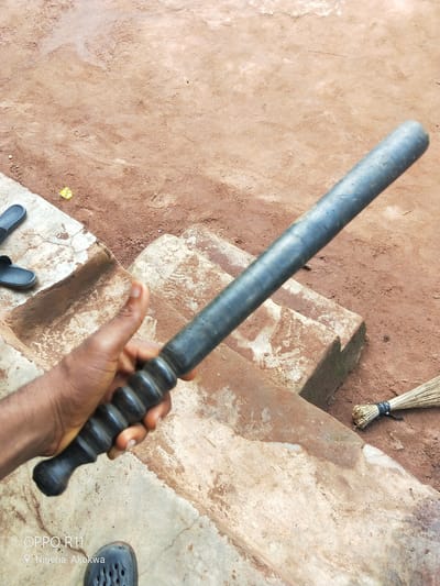 A person's hand holds a dark-colored baton with a ribbed handle. The baton is positioned diagonally across the frame, with the handle closest to the viewer and the tip extending upwards and to the right. The hand appears to be dark-skinned, and the fingernails are visible.
The background consists of a dirt ground with some texture, possibly indicating dried mud or footprints. To the left, there are concrete steps or ledges, with a pair of dark-colored flip-flops placed on the uppermost surface. Further to the right, a broom made of natural fibers is leaning against the edge of the concrete structure, its bristles fanned out on the dirt ground.
The scene appears to be outdoors, with natural lighting. The overall impression is one of a casual or possibly utilitarian setting. The colors are predominantly earthy tones of brown and gray, with the dark baton and flip-flops providing contrast.
Visible text at the bottom left of the image reads "OPPO R11" and "Nigeria Akokwa," indicating the phone model used for the photograph and the geographical location. There are no explicit signs of activity or event taking place, beyond the implied act of holding the baton. The time of day and weather conditions are not discernible.