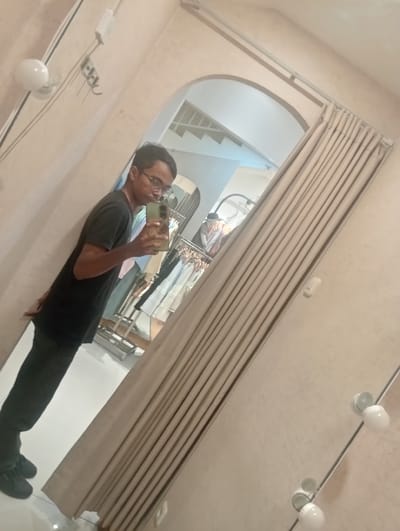 A person is taking a mirror selfie in what appears to be a fitting room or a boutique. The subject is wearing a black t-shirt, dark pants, and black sneakers. They have short, dark hair and are wearing glasses. The mirror reflects a rack of clothing behind the person, suggesting a retail environment. In the reflection, there are several garments hanging on hangers, mostly light-colored. The fitting room has a light beige or cream-colored wall and a large mirror that covers a significant portion of the wall. A beige curtain is drawn to the side, revealing a section of the mirror. The floor is a light, reflective tile. There are two white ceiling lights visible on the right side of the image, with another light fixture visible on the left. The overall lighting suggests it could be daytime or a well-lit indoor space. The person's pose and the selfie attempt indicate a casual and personal moment. The setting appears to be indoors, possibly a clothing store, in Larangan, Indonesia.