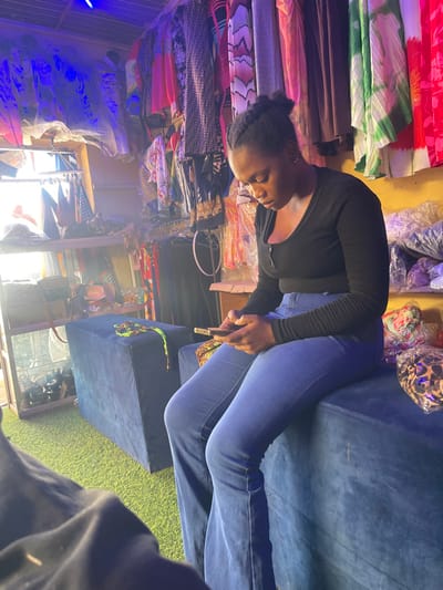 This image captures an interior scene within a retail clothing store or boutique in Samaru, Nigeria. A young adult female with dark skin and dark braided hair is prominently seated on a blue velvet ottoman, wearing a black long-sleeved top and blue flared jeans. She is focused on a smartphone held horizontally in both hands. The store's interior features walls lined with numerous garments of various colors and patterns, displayed on hangers and racks. Some merchandise, including what appears to be bags or accessories, is packaged in clear plastic and stacked. Shelving units on the left also hold various items. The floor is covered with green artificial turf. Overhead, blue LED strip lights illuminate parts of the space, while bright white light enters from an opening on the left side. An additional blue velvet ottoman is visible to the female's left, with a green and yellow beaded accessory resting on it.