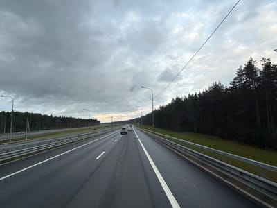 The image shows a view from a vehicle traveling on a highway in the city of Priladozhsky, Russia. The highway has multiple lanes, separated by white lines and guardrails. On the right side of the highway, there is a dense forest of pine trees. The sky is overcast with dark clouds, suggesting a cloudy or potentially rainy day. A few cars are visible on the highway, moving in the same direction as the viewer. Lampposts are present along the side of the road, indicating it is a developed highway. The overall scene depicts a typical highway landscape in a forested area under a cloudy sky.