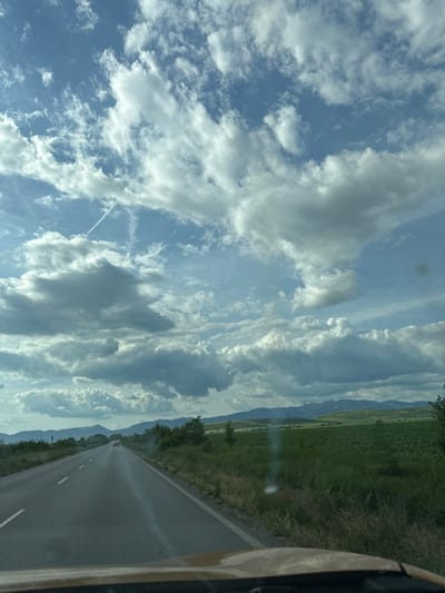The image shows a view from inside a vehicle, likely a car, traveling on a paved road. The road appears to be a rural highway with a double yellow center line and a single white line marking the edge of the road. The view is predominantly of the road ahead, framed by a field of grass and trees on the right and a cloudy sky with streaks of mountains in the distance. The car appears to be moving toward mountains in the distance, and there is a small town on the road ahead, which looks like it is Krushare, Bulgaria.