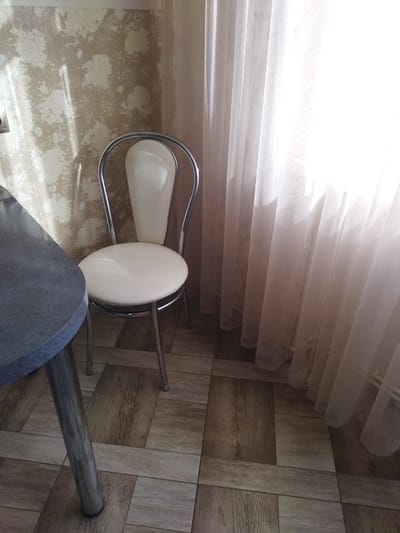 This is a photograph of an interior scene. In the foreground, a portion of a dark, speckled table is visible on the left side of the frame. To the right of the table, a chair with a chrome frame and a cream-colored padded seat and back is positioned. Behind the chair, sheer, light-colored curtains hang, obscuring a window. The walls appear to be covered in textured wallpaper with a beige and off-white pattern. The floor is tiled with a wood-grain pattern. The lighting suggests it is daytime, with natural light filtering through the curtains, creating bright highlights and shadows. The photograph was taken in Dolyna, Ukraine.