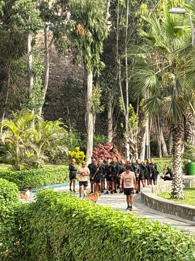The image depicts a line of individuals in the city of Ancón, Peru, within a park-like setting characterized by trees and manicured greenery. The group, comprised of at least a dozen people, is situated on a paved walkway or path. Many appear to be wearing matching attire consisting of dark-colored jackets or uniforms. A few individuals in the line are wearing civilian attire. A dog is observed near one of the people. The surrounding environment is lush with various tree species and dense bushes. A stone wall or border is seen along the path. The image is taken during daylight, with natural light illuminating the scene.
