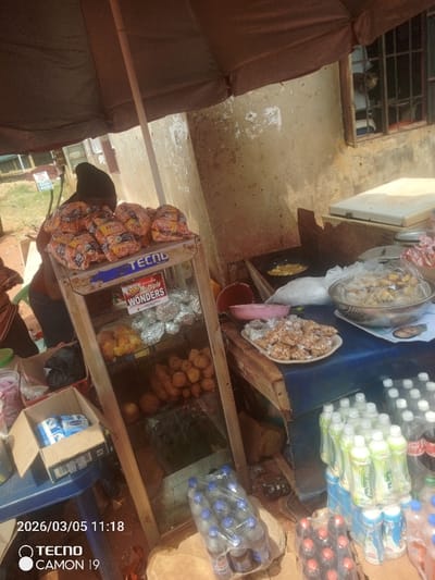 An outdoor food and beverage stall is operating under a large brown umbrella in Nsugbe, Nigeria. A person wearing a dark cap and an orange-brown top is partially visible on the left, positioned behind a counter, facing away from the camera.

The stall consists of a glass-fronted display case on the left, labeled "TECNO" at the top and featuring a "2016 Multiple WONDERS" sign inside. This case holds various baked goods, including foil-wrapped items, golden-brown buns, and spherical fried items. On top of the display case, several packages of "ROMA" branded bread are stacked.

To the right, a makeshift wooden counter or table, partially covered with a blue fabric, displays additional food items. A metal pan contains food cooking, possibly eggs or a similar fried item. Nearby are a pink bowl, a white plate holding small fried snacks (possibly chin-chin), and a metal colander filled with more fried snacks.

On the ground and on a blue plastic stool, multiple plastic-wrapped packs of bottled beverages are arranged for sale, including "Nutri-Milk" dairy drinks, "Pepsi" soft drinks, and other bottled liquids. A brown cardboard box containing white packets is also visible on the stool.

The background features a weathered, light-colored concrete wall with a window fitted with metal bars. The ground is exposed earth. The scene is illuminated by natural light, indicating daytime. The image was captured on 2026/03/05 at 11:18 with a TECNO CAMON 19 device.