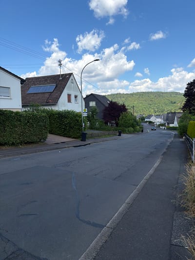 The image shows a street scene in Idar-Oberstein, Germany. A paved road runs diagonally across the frame, bordered by a curb and a grassy area with dry grass at the top edge. On the left side of the road, there are several houses with pitched roofs. One house in the foreground has solar panels installed on its roof. A tall streetlight with a curved arm stands on the left side of the road, near the houses.
A dark-colored car is visible on the road, driving away from the viewer. Further up the street, more houses are nestled on a hillside covered in lush green trees. The sky above is a bright blue with scattered white clouds. The scene appears to be set during the daytime, likely late morning or early afternoon, given the bright sunlight and clear skies. The overall atmosphere is peaceful and suburban, with residential buildings and a natural, tree-covered landscape forming the backdrop. There are no people visible in the image. A bus stop sign with the letter "H" in a yellow circle is visible on a pole next to a black trash bin, adjacent to the road.