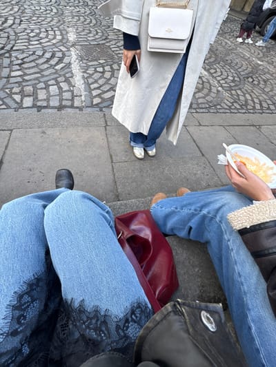 Three people are seated outdoors on a concrete surface. One person wears a light-colored trench coat, jeans, and metallic sneakers, holding a phone. Another person wears a shearling-lined coat, jeans, and tan shoes, holding a plate with food. The third person wears jeans with black lace trim and black shoes, with a red purse and a black bag nearby. The surface under them is a mix of concrete and cobblestones.