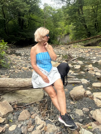 A Caucasian woman with short blonde hair and sunglasses sits on a fallen log near a rocky stream in a wooded area. She is wearing a blue and white polka dot strapless top and a white skirt, with black and pink athletic shoes. A black backpack is placed behind her on the log. The stream bed is filled with stones and some water is visible. Trees and foliage surround the scene, with a visible landslide embankment in the background. The location is identified as Plastunka, Russia.