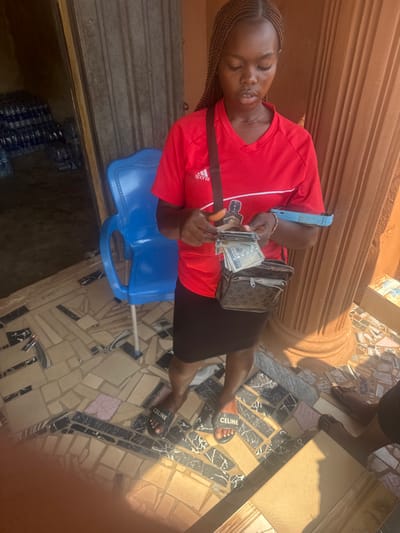 A female individual stands outdoors in Aguata, Nigeria, focusing on handling currency. She is seen from the chest up, with her head slightly bowed, concentrating on a stack of Nigerian Naira banknotes in her hands, including a visible 1000 Naira note. She wears a red Adidas short-sleeved shirt, a black skirt, and black "CELINE" branded slide sandals. A dark brown shoulder bag with a repeating pattern is slung across her body. A light blue mobile phone is held or tucked under her right arm. The ground beneath her is a mosaic-style tiled surface composed of irregular beige, black, and white pieces. To her left, a blue plastic chair sits near an open dark brown doorway, beyond which numerous stacks of bottled water are visible, suggesting a retail or storage space. To her right, a light brown fluted architectural pillar is present. The lower legs and feet of a second individual, wearing dark flip-flops, are partially visible in the far right foreground. The scene is illuminated by bright natural light, casting distinct shadows.