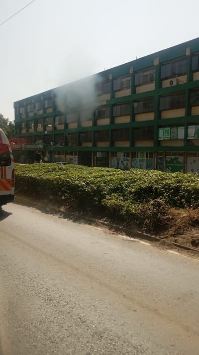 A three-story green building, labeled "Matata House," emits smoke from a section. A Cooperative Bank sign is visible on the ground floor. A dense green hedge separates the building from the road. The road, gray and slightly worn, occupies the foreground of the frame. A vehicle, partially visible on the left, suggests the photo was taken from within another car. The sky is bright and hazy.