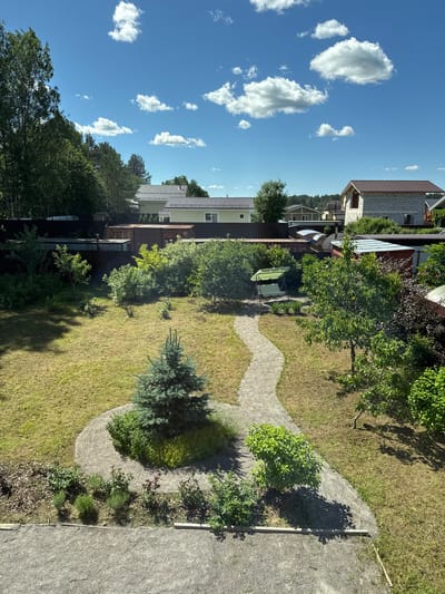 This media file depicts a sunny day in a suburban backyard in Steklyannyi, Russia. The scene is captured from an elevated perspective, looking down at a lush green lawn interspersed with trees, bushes, and a winding gravel pathway. A light green swing set is visible near a tree.
Several houses with pitched roofs line the background, suggesting a residential neighborhood. The sky is a vibrant blue with scattered, fluffy white clouds. The time of day appears to be midday, given the bright sunlight and the distinct shadows cast by the trees.
The overall impression is one of a peaceful, well-maintained outdoor space during pleasant weather. No people are visible in the image. There is no visible text in the media file.