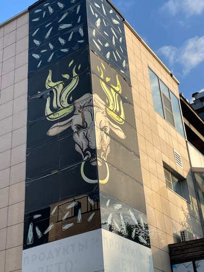 This is an eye-level, medium shot of a building in Ivanovskaya, Russia, with a large mural of a bull's head on the corner. The building is clad in beige tiles and has several windows. The mural is black and yellow, with the bull's head depicted in detail, including its horns and a ring in its nose. The mural also has white scribbled lines on a black background above the bull's head and white text on a white background at the bottom. The sky is blue with some clouds.