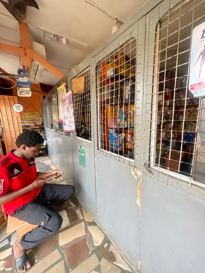In Akowonjo, Nigeria, a young Black man, wearing a red Adidas shirt with black stripes and black pants, sits on the floor outside a retail establishment. He is holding a smartphone in his hands and appears to be looking at it. The retail establishment has a grey metal security gate with a checkered pattern that covers the windows. Inside the store, shelves are stocked with various products, including bottles and boxes of what appear to be medications or toiletries. A sign with a picture of a person on it is visible on the outside of the store, and other posters are attached to the wall near the gate. The floor is tiled with irregular-shaped stones in shades of brown and beige. The surrounding environment includes a concrete ceiling and orange-colored wooden structures.