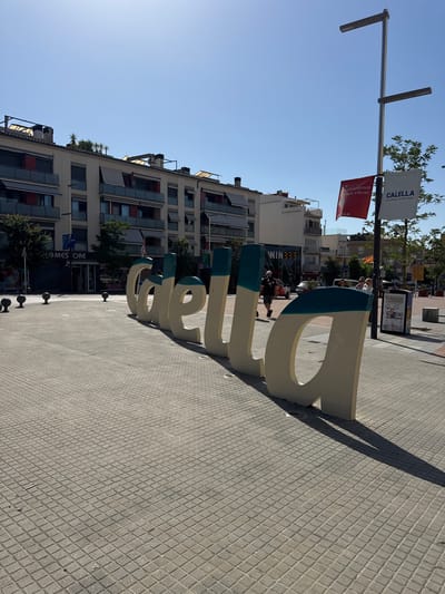 The image depicts a sunny outdoor scene in the city of Calella, Spain. In the foreground, large, three-dimensional letters spelling out "CALELLA" are prominently displayed on a paved plaza. The letters are painted in a light beige color with a teal stripe across the top. To the right of the letters, a tall lamppost with two lights is visible, from which two banners are hanging. One banner is red and features white text, while the other is white with blue text and reads "CALELLA" followed by "La ville qui te fait vibrer" in smaller print.
In the background, there are several multi-story buildings with balconies and awnings. A few trees are planted along the street. The plaza is paved with square tiles arranged in a diamond pattern. Several people are walking in the scene. One person, wearing a baseball cap and a t-shirt, walks towards the left of the frame past the letters. Another group of people is standing near a trash receptacle to the right. A store with a sign reading "HOMES STORE" is visible on the left side of the plaza. The sky is clear and blue.