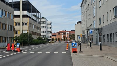 The image shows a street scene in Linköping, Sweden. In the foreground, there is a road with a pedestrian crossing marked by white squares. Orange traffic cones and a blue and white pedestrian crossing sign are positioned on the right side of the road. On the left side of the road, there are more orange traffic cones, one of which is a reflective barrier.
The buildings flanking the road are modern, multi-story structures. The building on the left is gray with yellow accents and rows of windows. The building on the right is a long, light gray building with a dark gray base and numerous windows. Further down the street, there are other buildings, including a red brick building with arched windows and a lighter-colored modern building with a white facade.
The sky is partly cloudy, and the overall lighting suggests it is daytime. There are some trees and greenery visible along the sides of the road. A few cars are present on the road, and a person can be seen sitting on a bench in the distance on the left. There is also a parking sign with a "P" symbol and "10 min" text visible on the right.