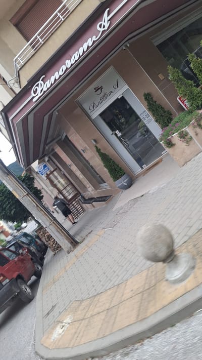 The image shows the exterior of a cafe named "Panorama A" located in Gotse Delchev, Bulgaria. The cafe has a prominent sign with the name "Panorama A" in white cursive lettering. The entrance to the cafe is a glass door with "Cafe Panorama A" written on it, along with what appears to be operating hours. There are two small green conical trees planted in large grey pots outside the cafe. A red car and a dark colored car are parked on the street in front of the cafe. A person wearing a black jacket and shorts is walking on the sidewalk away from the cafe. The sidewalk is paved with interlocking bricks and has a yellow-colored border. A concrete sphere on a pedestal is visible on the sidewalk. The building facade is made of beige-colored tiles. The overall scene depicts a street view in front of a business.